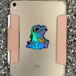 K&ouml;nnte beinhalten: Ein iPad Air in einer transparenten H&uuml;lle mit einem rosa Stifthalter. Auf der R&uuml;ckseite befindet sich ein holografischer Aufkleber eines Zeichentrickkrokodils mit roten Augen, einem gelben Kragen und einem roten R&uuml;ckgrat.
