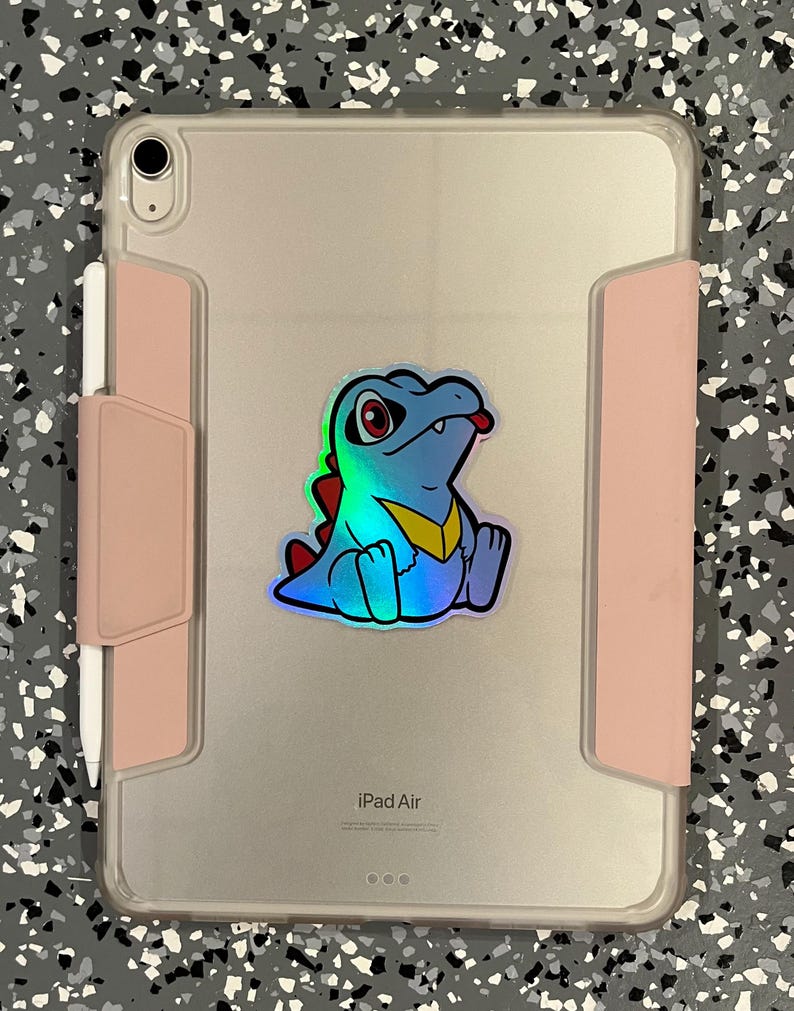 K&ouml;nnte beinhalten: Ein iPad Air in einer transparenten H&uuml;lle mit einem rosa Stifthalter. Auf der R&uuml;ckseite befindet sich ein holografischer Aufkleber eines Zeichentrickkrokodils mit roten Augen, einem gelben Kragen und einem roten R&uuml;ckgrat.