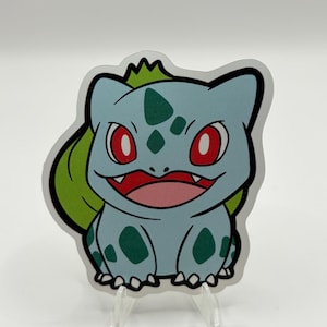 May include: A sticker featuring a cartoon Bulbasaur character. The Bulbasaur has a light blue body with green spots, red eyes, and a green bulb on its back. The sticker is outlined in black and is displayed on a clear stand.