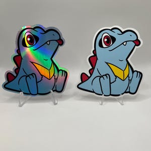 K&ouml;nnte beinhalten: Zwei Aufkleber eines Zeichentrick-Dinosauriers mit blauen K&ouml;rpern, roten R&uuml;ckenstacheln und gelben Brustmarkierungen. Ein Aufkleber hat ein holografisches Finish, der andere ist matt. Beide haben rote Augen und ihre Zungen schauen heraus. Sie stehen auf transparenten Kunststoffst&auml;ndern.