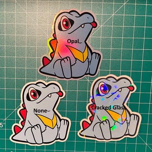 K&ouml;nnte beinhalten: Drei Aufkleber mit einem Zeichentrick-Dinosaurier mit roten Augen, einer gelben Brust und einer roten Zunge. Die Aufkleber tragen die Aufschriften "Opal", "None" und "Cracked Glas".