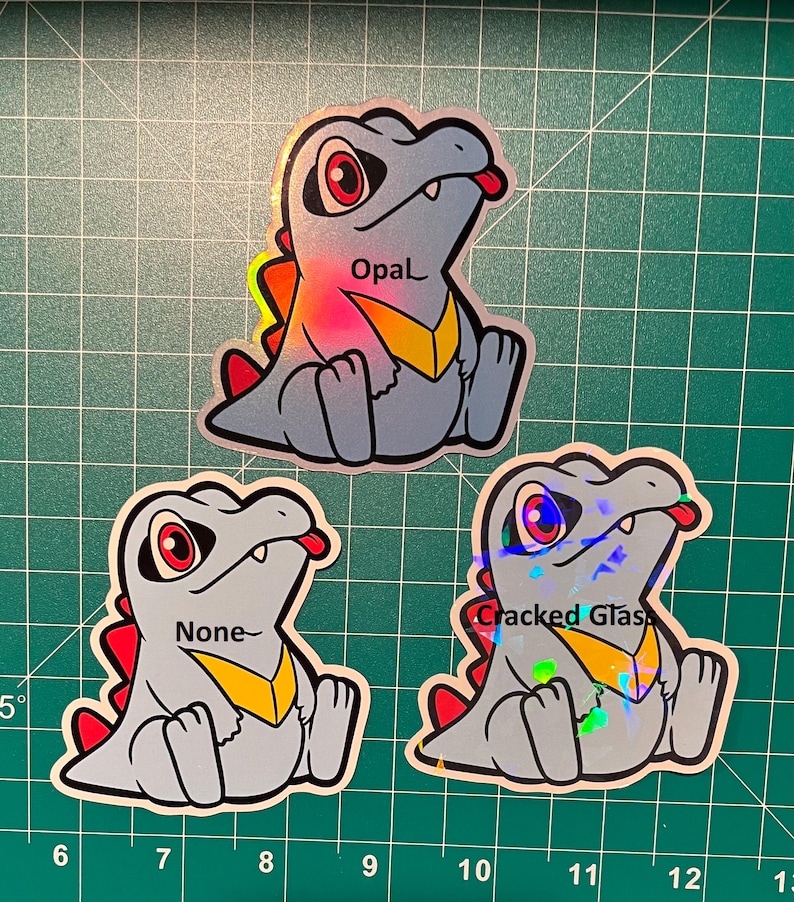 K&ouml;nnte beinhalten: Drei Aufkleber mit einem Zeichentrick-Dinosaurier mit roten Augen, einer gelben Brust und einer roten Zunge. Die Aufkleber tragen die Aufschriften "Opal", "None" und "Cracked Glas".