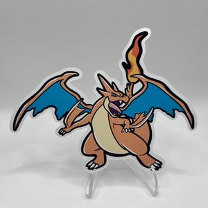 May include: A stylized, orange and cream-colored Charizard figure with blue wings and a fiery tail. The figure is mounted on clear plastic supports, showcasing a dynamic pose. The artwork features bold black outlines and is a collectible item.