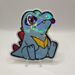 K&ouml;nnte beinhalten: Ein holografischer Aufkleber mit einem Cartoon-Krokodil mit blauem K&ouml;rper, roten Stacheln und gelber Brust. Das Krokodil hat eine rote Zunge, die herausragt, und sitzt. Der Aufkleber wird auf einem transparenten St&auml;nder ausgestellt.