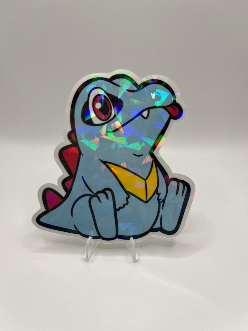 K&ouml;nnte beinhalten: Ein holografischer Aufkleber mit einem Cartoon-Krokodil mit blauem K&ouml;rper, roten Stacheln und gelber Brust. Das Krokodil hat eine rote Zunge, die herausragt, und sitzt. Der Aufkleber wird auf einem transparenten St&auml;nder ausgestellt.