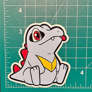 K&ouml;nnte beinhalten: Ein Aufkleber mit einer cartoonartigen, alligator&auml;hnlichen Kreatur mit grauem K&ouml;rper, roten Augen und roter Zunge. Es hat eine gelbe Brust und rote Stacheln. Der Aufkleber befindet sich auf einem gr&uuml;nen Rasterhintergrund.