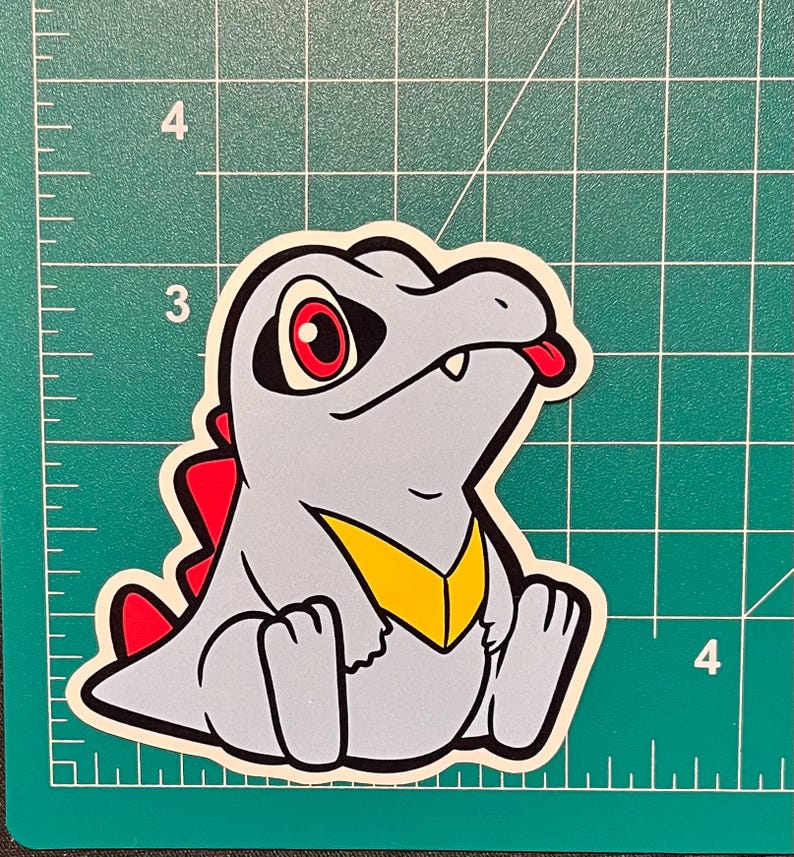 K&ouml;nnte beinhalten: Ein Aufkleber mit einer cartoonartigen, alligator&auml;hnlichen Kreatur mit grauem K&ouml;rper, roten Augen und roter Zunge. Es hat eine gelbe Brust und rote Stacheln. Der Aufkleber befindet sich auf einem gr&uuml;nen Rasterhintergrund.