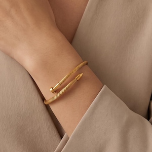 Op de afbeelding: Een goudkleurige metalen armband met een gedraaide kabelstructuur. De armband heeft een open ontwerp met twee uiteinden in de vorm van een spijkerkop en een puntige spijkerpunt, wat zorgt voor een wikkelend effect rond de pols.