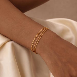 May include: A set of three thin, gold-toned bangles worn on the wrist. Each bangle features a delicate, textured surface with a repeating pattern of small, horizontal indentations that catch the light, creating a subtle shimmer across the metal.