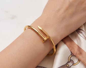May include: A gold-toned metal bracelet shaped like a bent nail, featuring a flat, circular head with small indentations and a pointed end that overlaps the main band, creating a continuous, open-ended circular design around the wrist.
