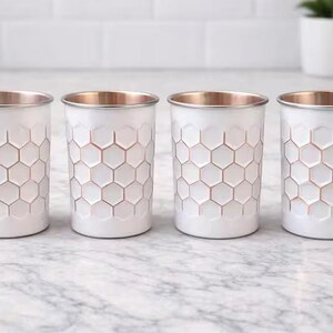 May include: Four white tumblers with a honeycomb pattern and copper-coloured rims. The glasses are arranged on a marble surface, offering a modern and stylish aesthetic. The design is geometric and visually appealing.