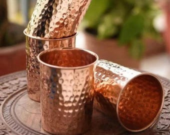 Puede incluir: Cuatro vasos de color cobre con una textura martillada. Tres vasos están apilados y uno está tumbado de lado. Los vasos están sobre una superficie de madera con detalles tallados. Los vasos tienen una superficie brillante y reflectante.