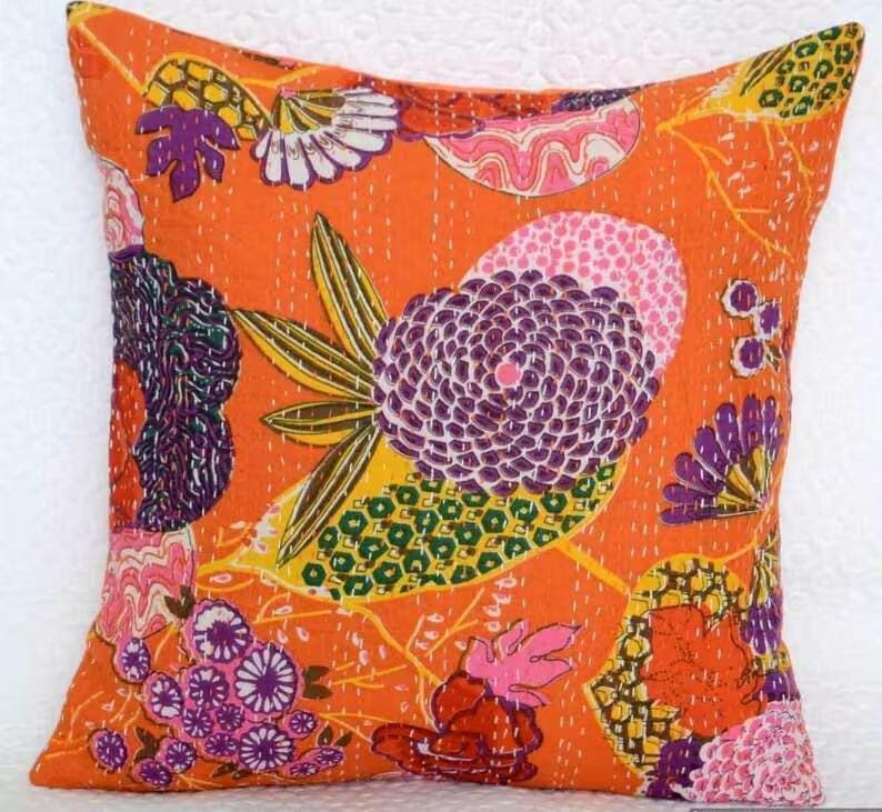 May include: Square orange pillow with a floral design. The pillow features a vibrant pattern of purple, pink, yellow, and green flowers and leaves. The pillow has a stitched texture and a solid orange background.