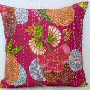 May include: A square decorative pillow with a vibrant pink background and a colorful floral pattern. The design includes orange, green, white, and blue elements, with intricate stitching details. The pillow is a decorative home accent.