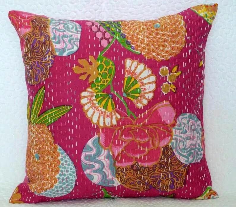 May include: A square decorative pillow with a vibrant pink background and a colorful floral pattern. The design includes orange, green, white, and blue elements, with intricate stitching details. The pillow is a decorative home accent.