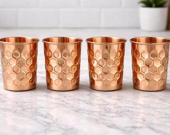 Puede incluir: Cuatro vasos de color cobre con un patrón de panal. Los vasos son cilíndricos con un borde ligeramente acampanado. Están dispuestos en fila sobre una superficie con vetas de mármol.