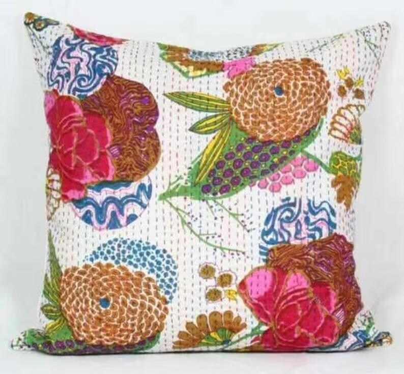 May include: Square throw pillow with a white background and a colorful floral pattern. The design features large flowers in shades of pink, brown, and orange, with green leaves and blue accents. The pillow has a stitched texture.