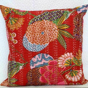 May include: Square red decorative pillow with a floral pattern. The pillow features a large orange flower, blue and pink accents, and green leaves. White stitching outlines the design.
