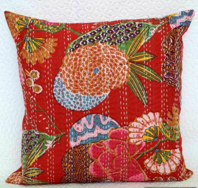 May include: Square red decorative pillow with a floral pattern. The pillow features a large orange flower, blue and pink accents, and green leaves. White stitching outlines the design.