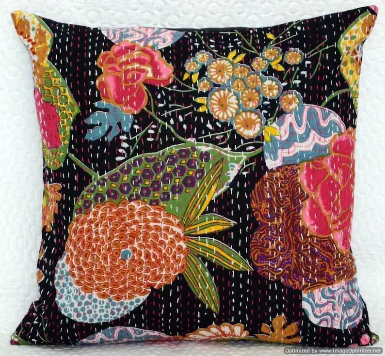 May include: Square decorative pillow with a black background and a colorful floral pattern. The design features large flowers in shades of pink, orange, and yellow, with green leaves and blue accents. The pillow is embellished with white stitching.