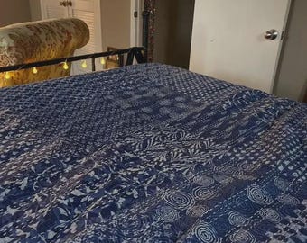 Puede incluir: Un edredón azul y blanco estampado cubre una cama. El edredón presenta varios diseños, incluidos patrones florales y geométricos. La cama está en una habitación con una puerta blanca y un sofá estampado.
