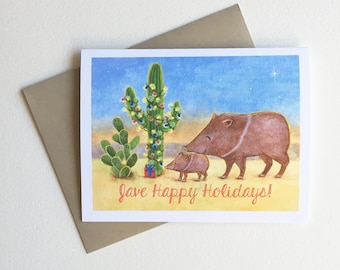 May include: A Christmas card featuring a cartoon illustration of a family of javelinas standing in front of a cactus decorated with Christmas lights. The card has the text "Have Happy Holidays!" in red.