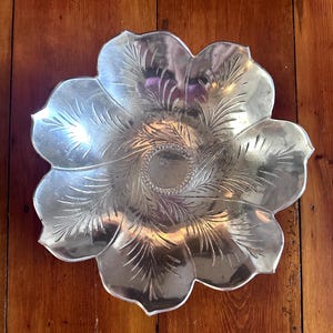 Op de afbeelding: Een zilverkleurige, bloemvormige decoratieve schaal. De schaal heeft zes bloemblaadjes met geëtste, veerachtige ontwerpen die vanuit het midden uitstralen. Het midden van de schaal heeft een cirkelvormig, gestructureerd patroon. De schaal staat op een houten oppervlak.