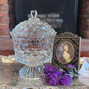 Puede incluir: Un bombonera de cristal transparente con tapa, colocada sobre una bandeja plateada. El plato tiene un patrón texturizado en forma de diamante. Un pequeño retrato enmarcado y flores moradas también están en la bandeja.