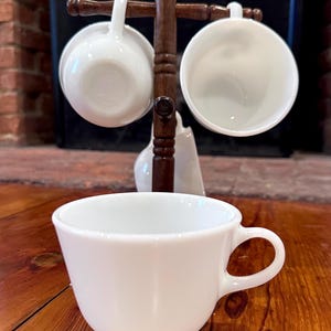 May include: A white ceramic teacup with a curved handle sits on a wooden surface. Two white teacups hang from a dark brown wooden rack. A small white creamer is also visible.