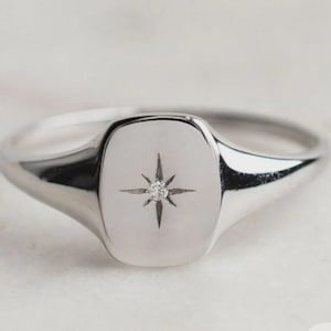 May include: A silver signet ring with a polished finish. The ring features a rectangular face with a starburst design and a small, clear gemstone at the center. The band tapers towards the back.