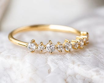 May include: A delicate gold ring featuring a series of small, sparkling diamonds. The ring's band is a thin, polished gold, and the diamonds are set in a row across the top. The ring is displayed on a white, textured surface.