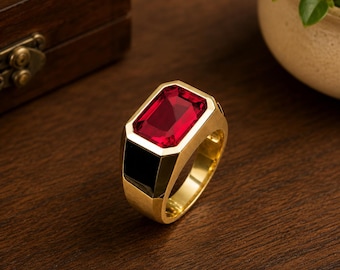 May include: A gold-colored ring with a large, rectangular, red gemstone. The ring has a black accent on the side and is set against a dark wooden surface. The ring is a statement piece.