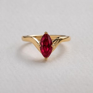 May include: A gold ring with a marquise-cut, deep red gemstone. The ring's band has a distinctive, curved design. The setting is a classic prong style, highlighting the vibrant colour of the stone. The ring is a piece of jewellery.