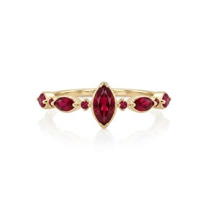May include: A gold ring featuring a central marquise-cut red gemstone flanked by two smaller marquise-cut red gemstones on each side, with tiny round red accent stones placed between them, all set in a polished yellow gold band.