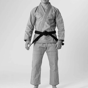 Pode incluir: Um uniforme de artes marciais cinza claro, ou gi, com uma faixa preta amarrada na cintura. O gi consiste em uma jaqueta e calças, com detalhes de costura visíveis. A pessoa está em pé em um fundo branco.