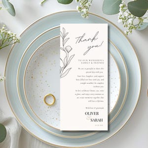 Könnte beinhalten: Eine Hochzeitstafel mit goldenem Ring, hellblauen Tellern und einer weißen Karte. Die Karte trägt die Aufschrift "Danke!" und enthält ein Blumendesign und eine Nachricht an Familie und Freunde.