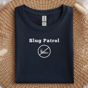 May include: Navy blue crew neck sweatshirt with the text "Slug Patrol" above a white graphic of a slug with a slash through it. The sweatshirt is folded on a woven surface.