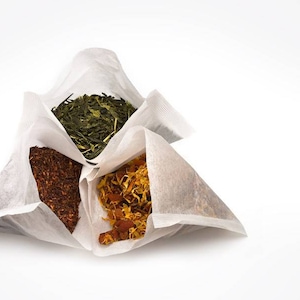May include: Four individual tea bags filled with different types of tea leaves. The tea bags are made of white paper and are folded into a pyramid shape. The tea leaves are visible through the paper. The tea leaves are green, brown, and yellow.