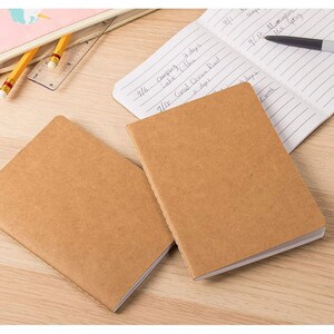May include: Two brown kraft paper notebooks with white lined pages. The notebooks are closed and stacked on top of each other. The top notebook has a white page showing with handwritten notes.