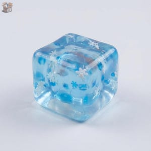 May include: A clear, blue resin cube with embedded white snowflake and blue dot designs. The cube is a solid, geometric shape, perfect for decorative purposes or as a unique game die. The cube is set against a plain white background.