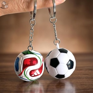 Puede incluir: Dos llaveros de balón de fútbol. Uno presenta un diseño blanco, rojo, azul y verde. El otro es un diseño clásico de balón de fútbol en blanco y negro. Ambos tienen anillos y cadenas de metal plateado.