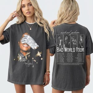 Puede incluir: Camiseta gris oscuro con un gráfico de Michael Jackson, un guante brillante cubriendo sus ojos. La parte trasera de la camiseta muestra una foto del concierto y el texto "BAD WORLD TOUR".