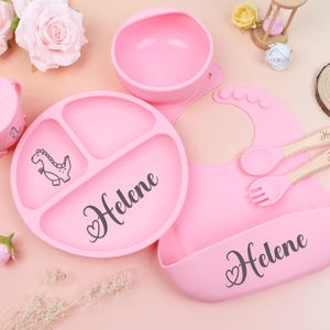 May include: A pink baby feeding set includes a divided plate with a dinosaur graphic, a bowl, a sippy cup, a bib, and utensils. The plate and bib are personalized with the name "Helene".