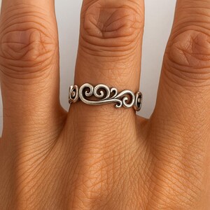 Puede incluir: Un anillo de plata con un delicado diseño de volutas. El anillo presenta un patrón continuo de rizos y bucles, creando un aspecto elegante y ornamentado. El anillo se lleva en un dedo, mostrando sus intrincados detalles y artesanía.