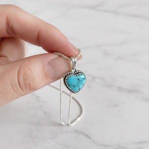 May include: A silver necklace with a heart-shaped pendant. The pendant has a turquoise stone and a silver rope-style border. The necklace is shown against a white marble background.
