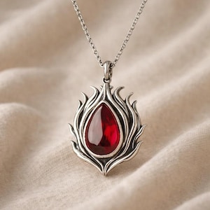 May include: A silver pendant necklace with a teardrop-shaped, deep red gemstone. The gemstone is set within a stylized flame design. The necklace hangs on a delicate silver chain, creating an elegant piece of jewellery.