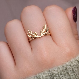 May include: A gold-colored ring featuring an antler design. The ring is worn on a finger, showcasing the delicate details of the antlers and the beaded band. The ring is a piece of jewelry.