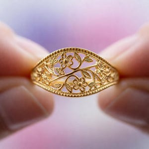 May include: A gold-colored ring with a floral filigree design. The ring features delicate flowers and leaves, creating an openwork pattern. The ring is held between two hands, showcasing the intricate details of the jewelry.