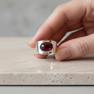 May include: A wide silver ring with a large, oval, deep red gemstone. The ring is held against a neutral background, highlighting the polished metal and the rich colour of the stone. The ring's design is simple but elegant.