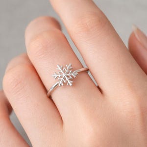 Puede incluir: Un anillo plateado con un diseño de copo de nieve. El anillo tiene una banda delgada, y el copo de nieve es el punto focal, con intrincados detalles calados. El anillo se lleva en un dedo, mostrando su delicada estética de temática invernal.